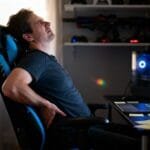 Ultra-realistic editorial photo of a remote worker captured mid-moment in a dim home office, leaning back slightly in a black-and-blue gaming chair with visible wear: loose stitching on the headrest, slight tilt in the frame, and a footrest partially extended. The worker adjusts their lower back with a tense expression, as if reacting to discomfort. A laptop glows on the desk beside scattered notes, hinting at long hours. Background shelves with blurred gaming gear emphasize the crossover between work and play. Shot in a candid mid-shot, hand-held documentary style with natural window light, slight motion blur on the worker’s hand, mild lens flare, and filmic grain ISO 1200. Background softly out of focus for realism, no stylization, no text.