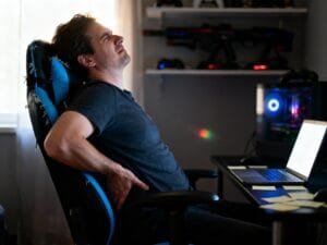 Ultra-realistic editorial photo of a remote worker captured mid-moment in a dim home office, leaning back slightly in a black-and-blue gaming chair with visible wear: loose stitching on the headrest, slight tilt in the frame, and a footrest partially extended. The worker adjusts their lower back with a tense expression, as if reacting to discomfort. A laptop glows on the desk beside scattered notes, hinting at long hours. Background shelves with blurred gaming gear emphasize the crossover between work and play. Shot in a candid mid-shot, hand-held documentary style with natural window light, slight motion blur on the worker’s hand, mild lens flare, and filmic grain ISO 1200. Background softly out of focus for realism, no stylization, no text.